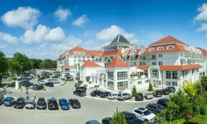 Elegancki hotel z białymi fasadami i czerwonymi dachami, otoczony zielenią i parkingiem pełnym samochodów.