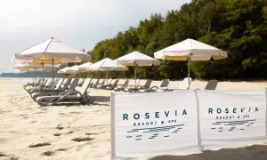 Plaża z leżakami i parasolami, w tle las, na pierwszym planie biały baner z logo "ROSEVIA RESORT & SPA".