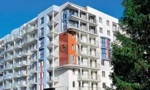 Nowoczesny hotel z balkonami i pomarańczowym akcentem architektonicznym, na tle błękitnego nieba i drzew.