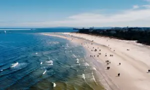 Plaża z ludźmi spacerującymi, morze i port z latarnią morską w tle.