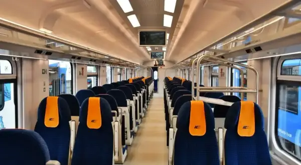 Wnętrze pustego wagonu kolejowego z rzędami niebieskich siedzeń i pomarańczowymi zagłówkami.