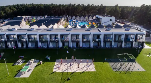 Widok z lotu ptaka na kompleks domków i budynków mieszkalnych z basenem i placem zabaw, na trawniku boisko do siatkówki i stoły do ping-ponga.