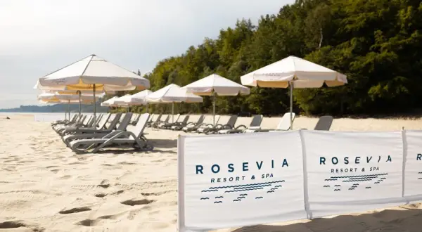 Plaża z leżakami i parasolami, w tle las, na pierwszym planie biały baner z logo "ROSEVIA RESORT & SPA".