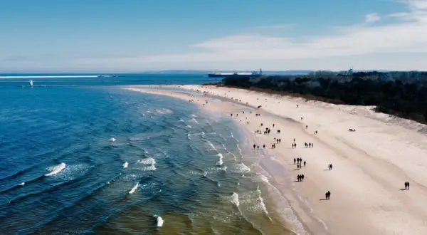 Plaża z ludźmi spacerującymi, morze i port z latarnią morską w tle.