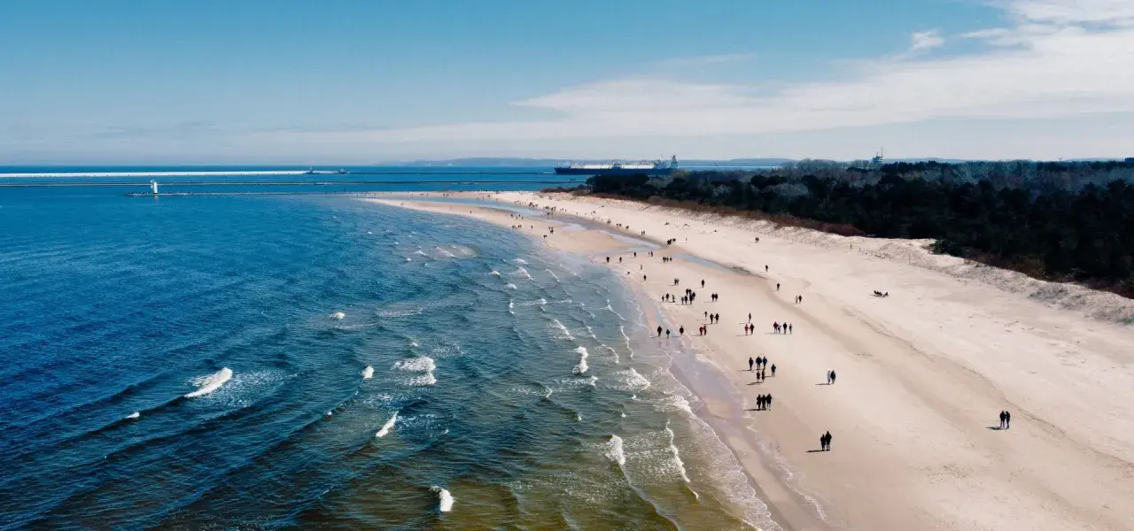 Plaża z ludźmi spacerującymi, morze i port z latarnią morską w tle.