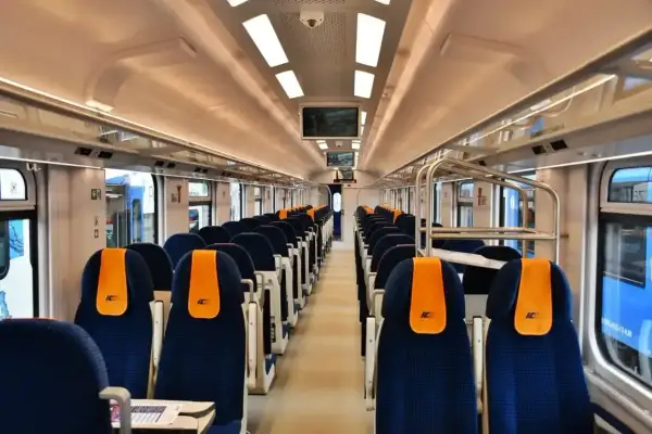 Wnętrze pustego wagonu kolejowego z rzędami niebieskich siedzeń i pomarańczowymi zagłówkami.