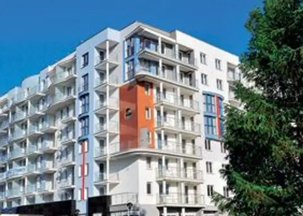 Nowoczesny hotel z balkonami i pomarańczowym akcentem architektonicznym, na tle błękitnego nieba i drzew.